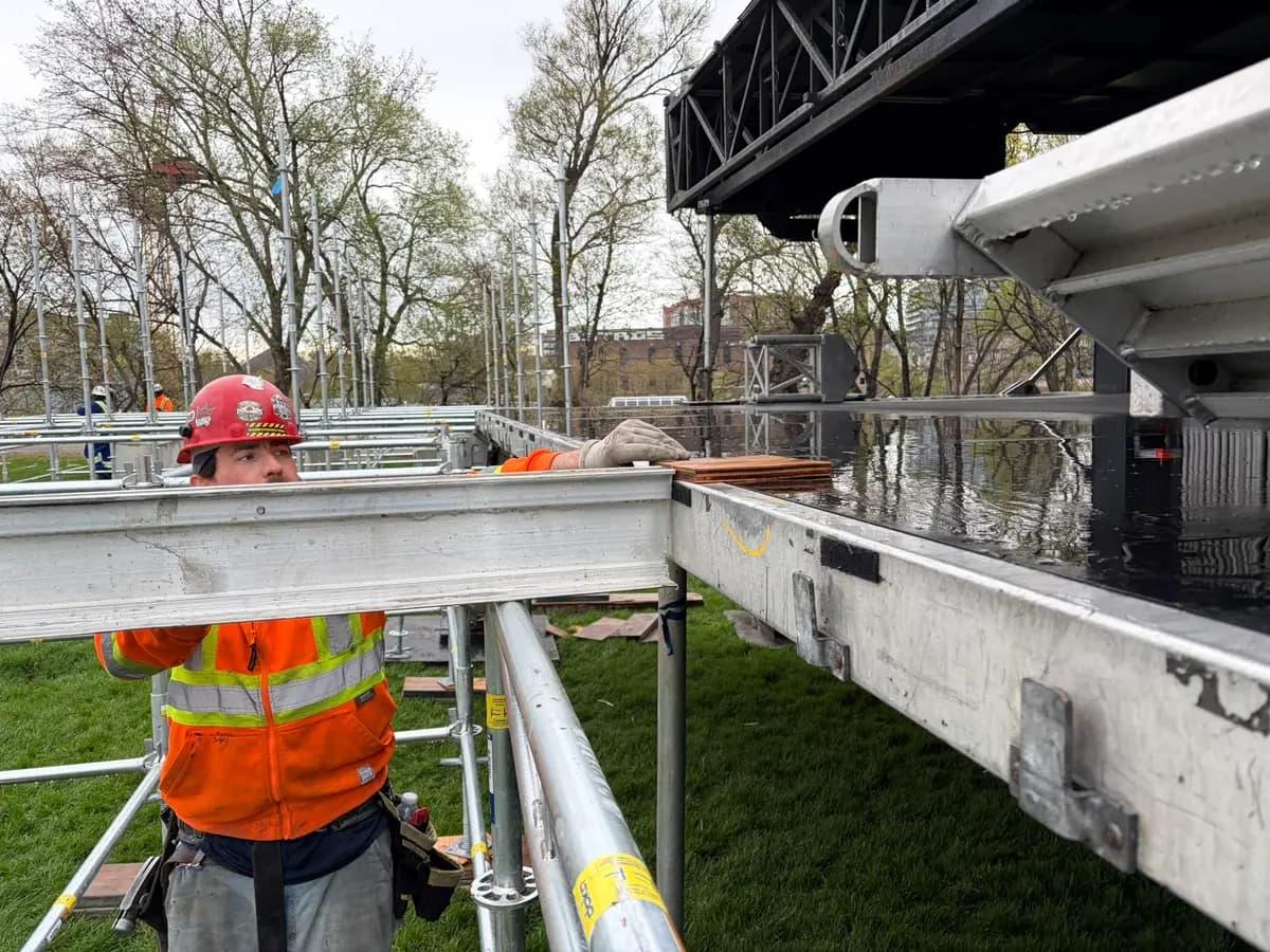 Crew member shimming and leveling a stage deck beam over scaffold shoring during an event build in a Toronto park