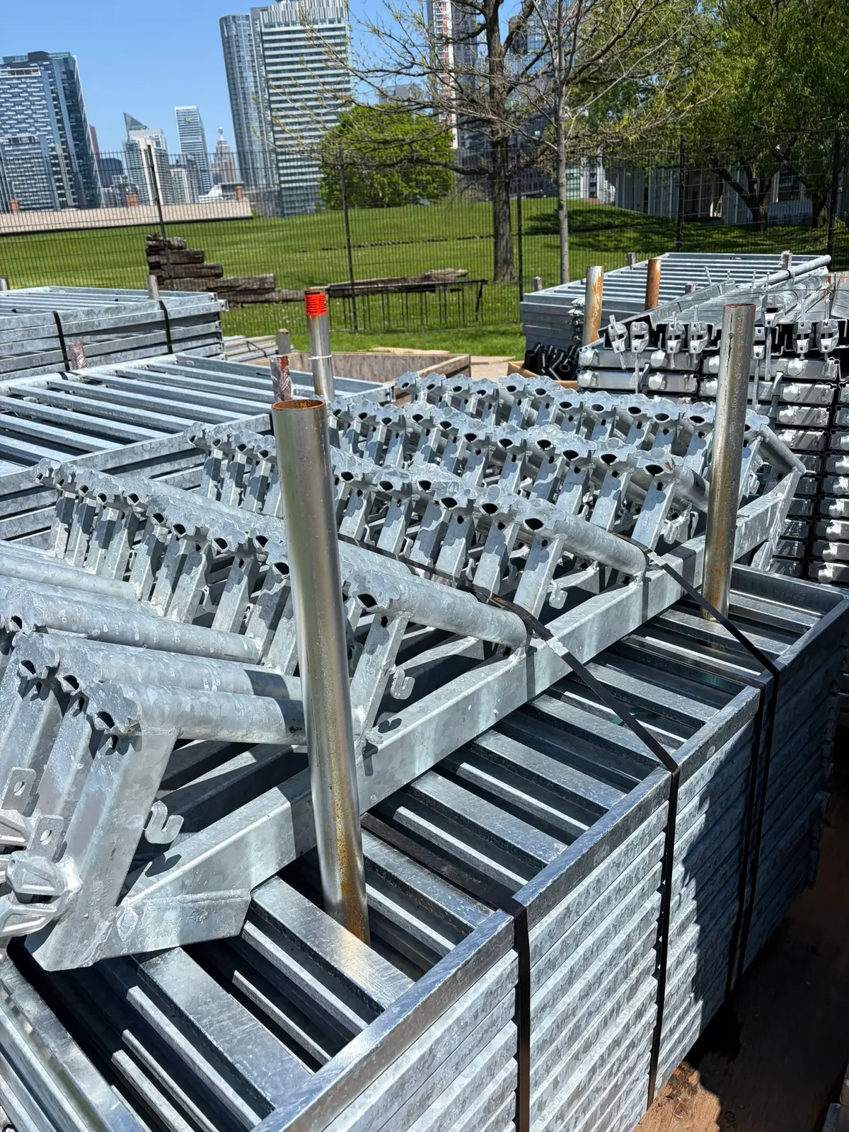 Pallets of galvanized scaffold frames and components staged in a laydown yard with the Toronto skyline behind.
