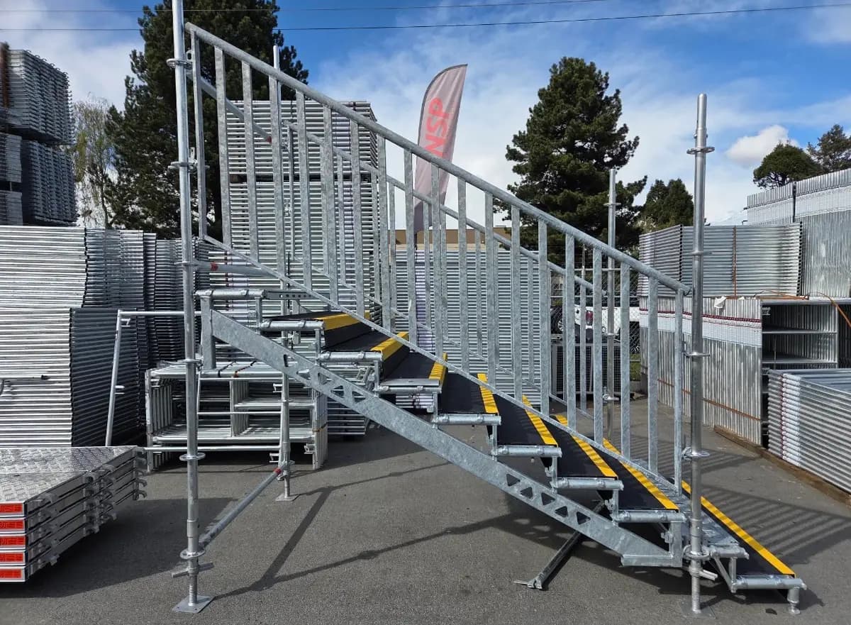 Galvanized scaffold stair unit with guardrails staged in an equipment yard beside stacked scaffold components