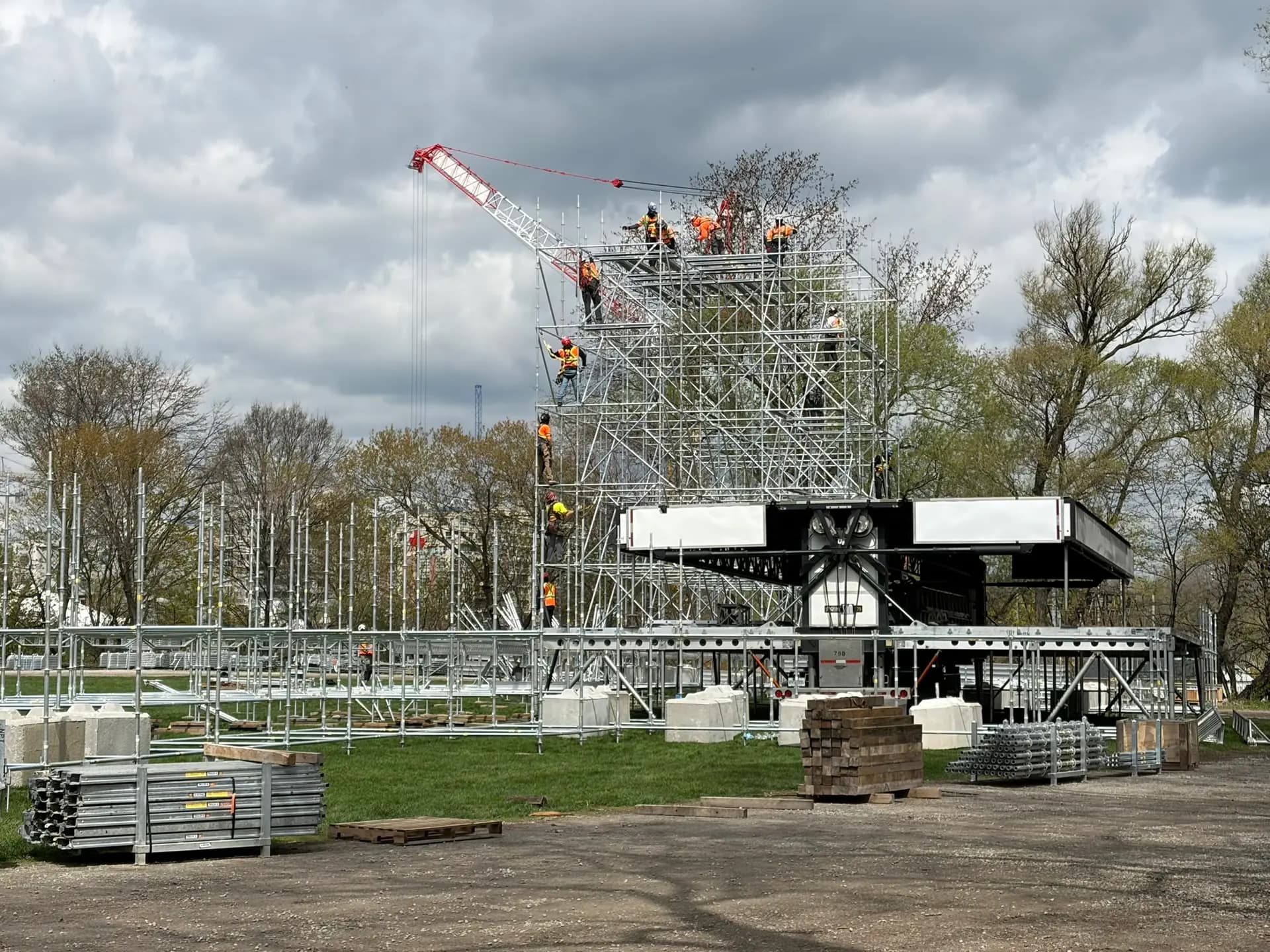 Mammoth crew erecting a festival stage scaffold tower at height under a dramatic sky, guided by a crane
