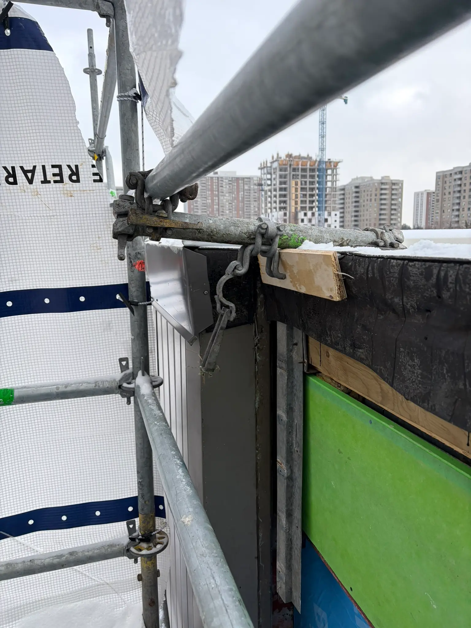 Scaffold guardrail and facade tie detail above a snowy jobsite with highrise construction in the background