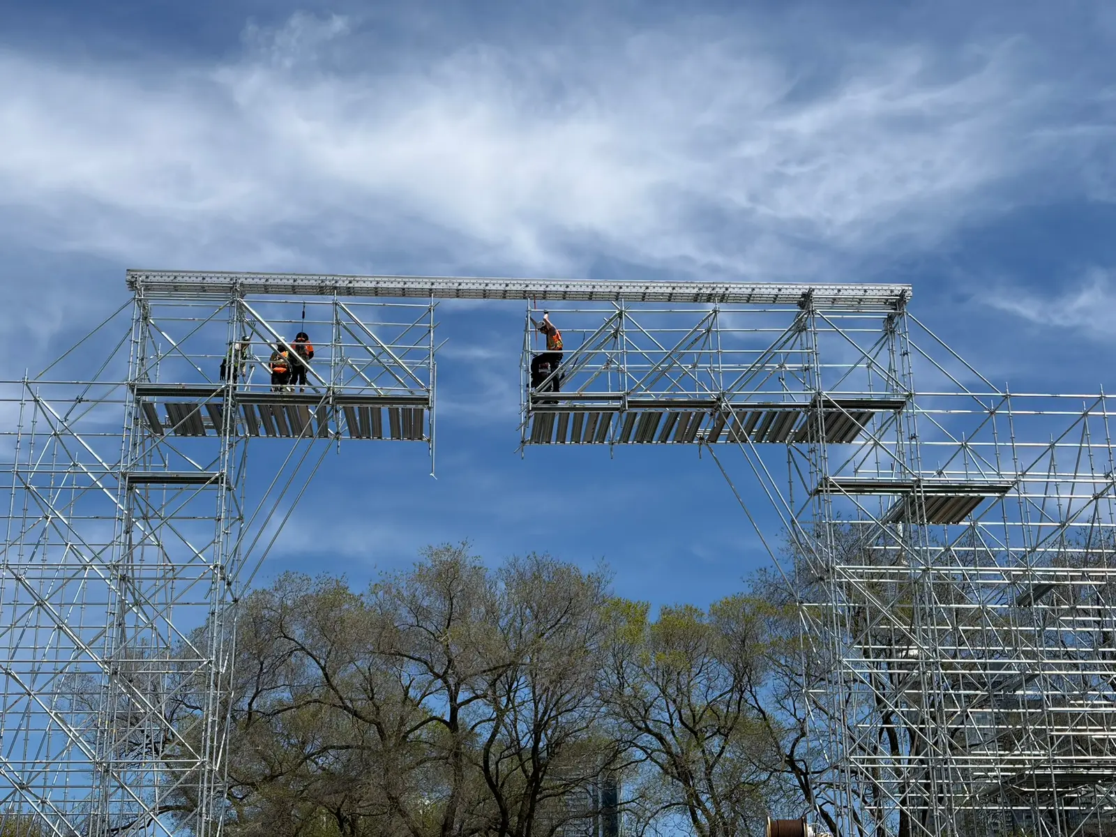 Scaffolders bridging two towers with a beam track high above the treeline during a festival build