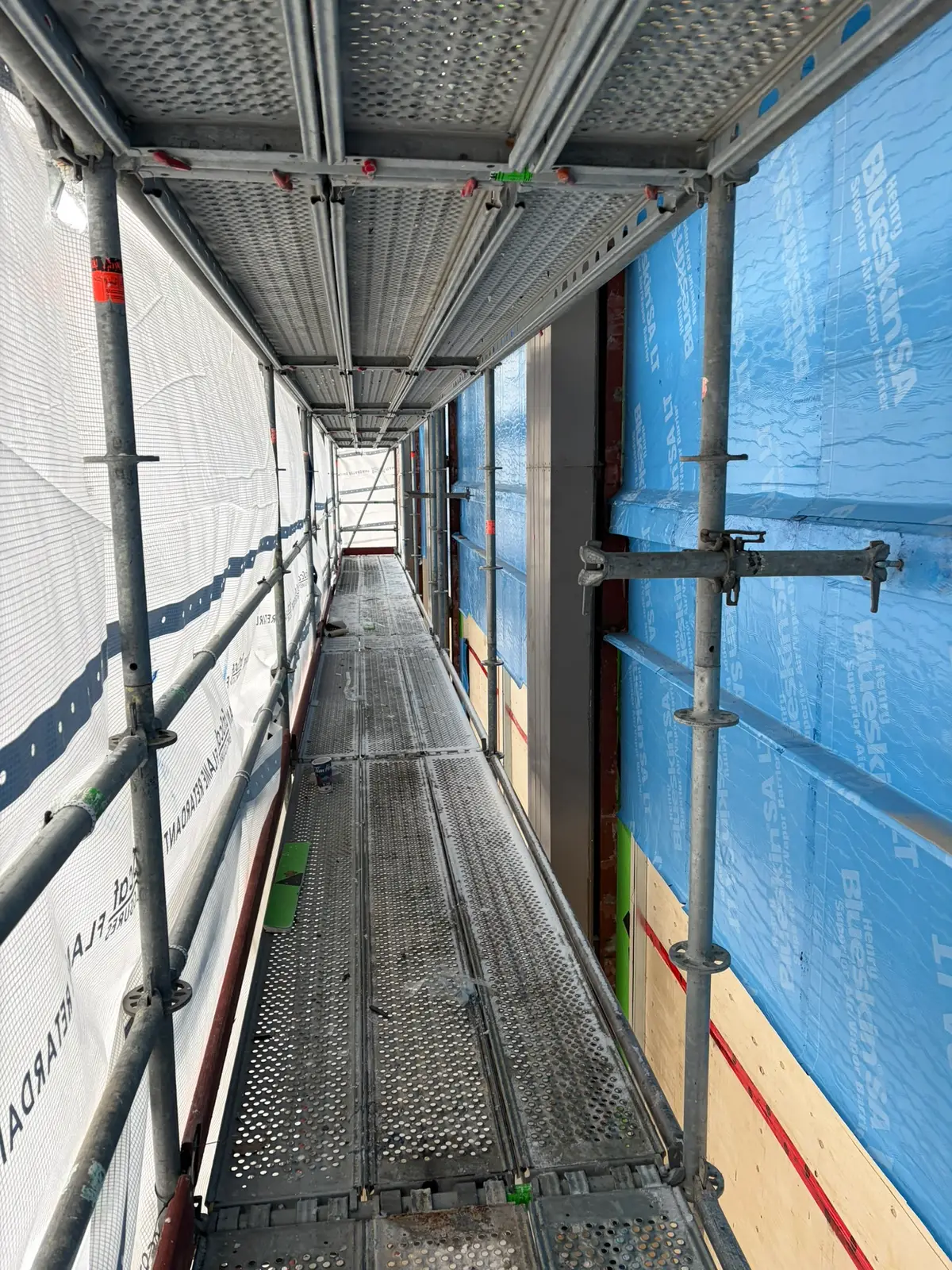 Long scaffold walkway with perforated steel decks and debris netting running along a membrane-wrapped facade