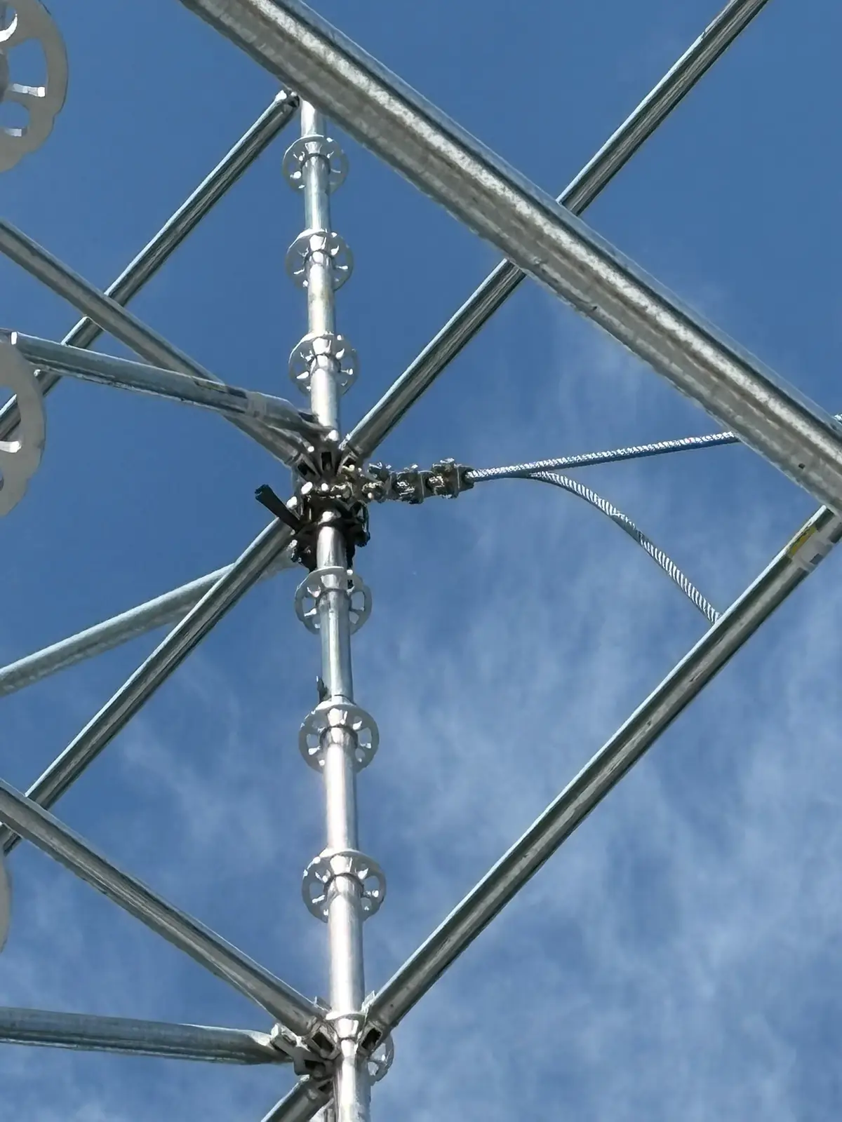 Ringlock scaffold standards with rosette connections and a guy wire seen from below against a blue sky