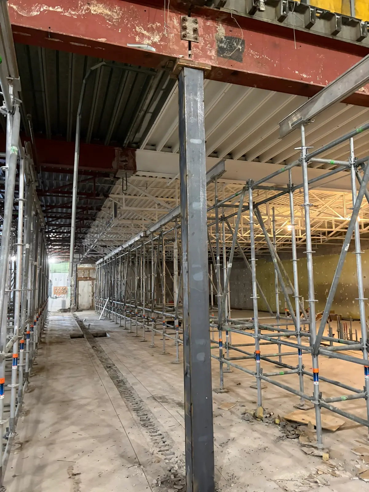 View down a warehouse aisle showing a new steel column and extensive shoring scaffold during structural upgrades