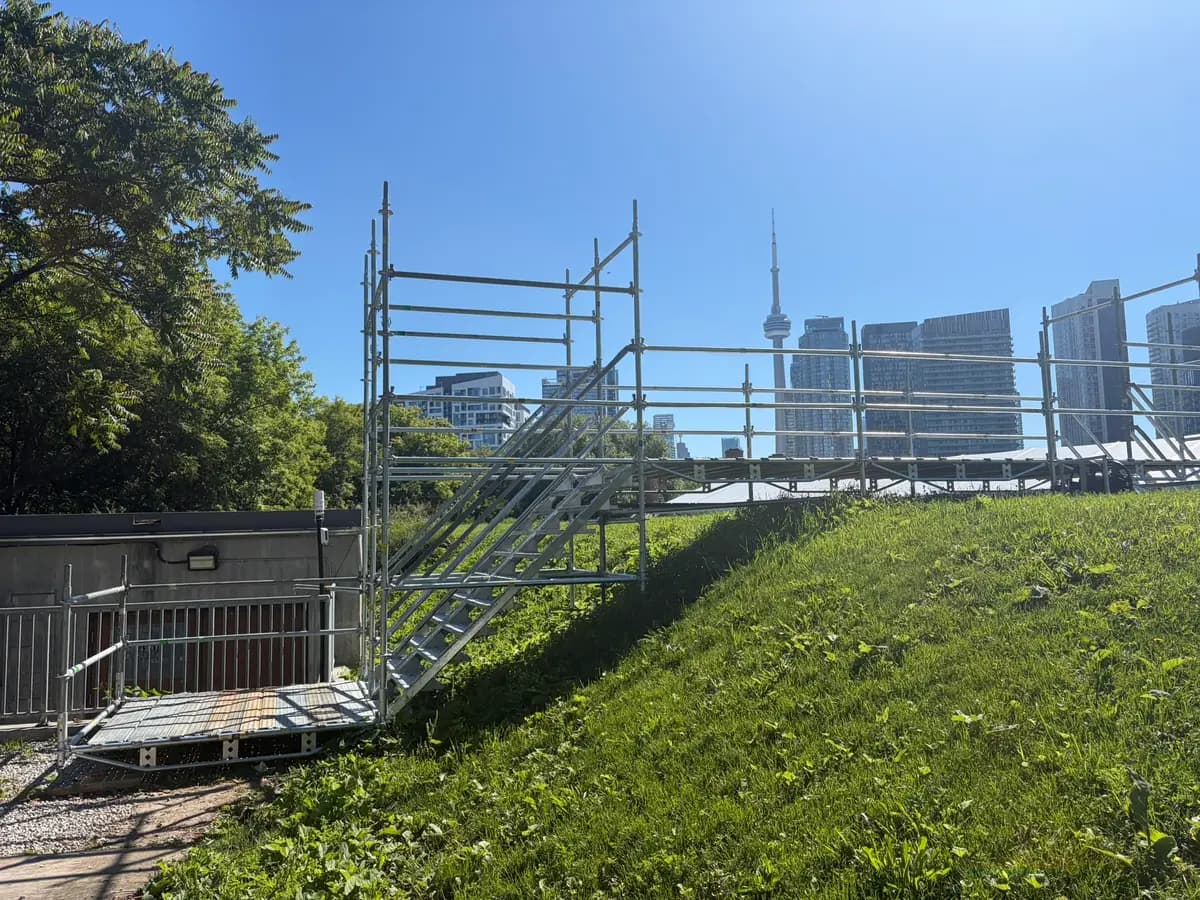 Galvanized scaffold stair and guardrail walkway crossing a grassy embankment with the CN Tower and Toronto skyline behind.